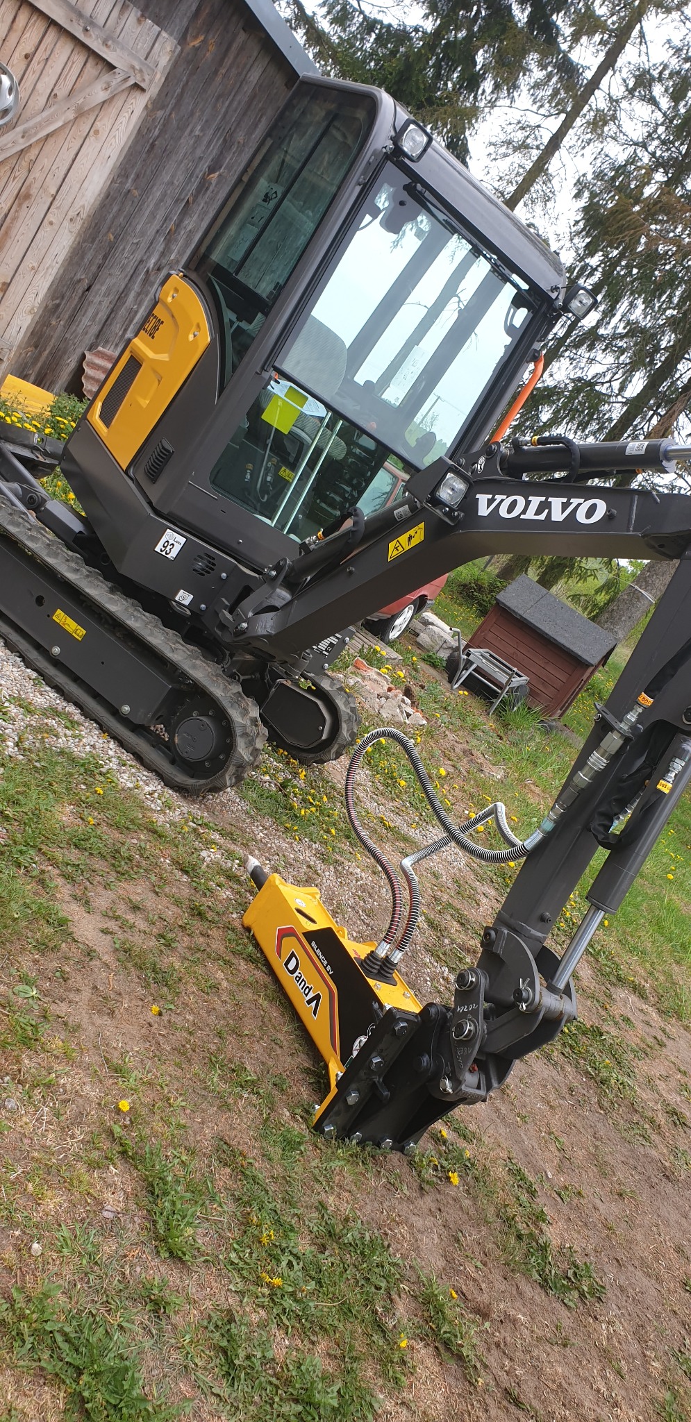 Żółto-czarny młot hydrauliczny Danda zamontowany na ramieniu minikoparki Volvo na terenie zielonym z dębową deską w tle.