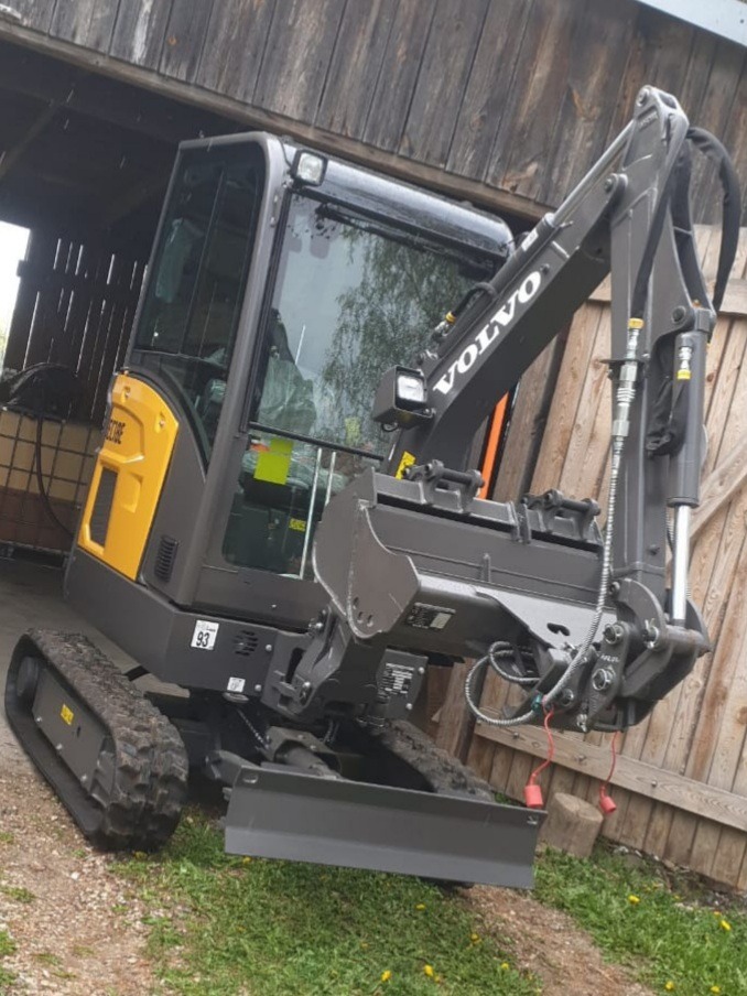 Nowa, kompaktowa koparka marki Volvo, zaparkowana na trawie przed drewnianym budynkiem gospodarczym, widok z boku, lekki kąt.