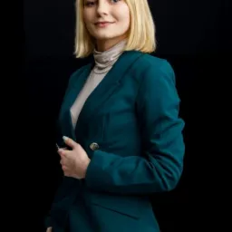 Portret kobiety w szmaragdowym garniturze na czarnym tle, z delikatnym uśmiechem i blond włosami do ramion. Profesjonalny wizerunek.