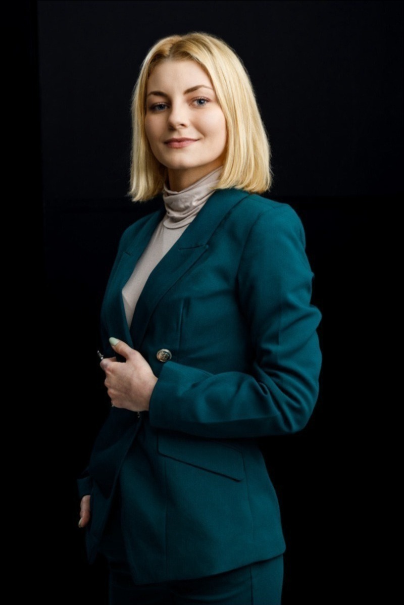 Portret kobiety w szmaragdowym garniturze na czarnym tle, z delikatnym uśmiechem i blond włosami do ramion. Profesjonalny wizerunek.