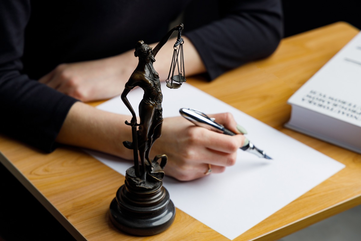 Statuetka Temidy na biurku, obok dłoni piszącej piórem na białej kartce. W tle książka, całość w ciepłej tonacji.