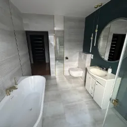 Nowoczesna łazienka z wanną wolnostojącą, umywalką z szafką, toaletą podwieszaną i dużym okrągłym lustrem, wykończona szarymi płytkami imitującymi kamień i ciemnozieloną ścianą.