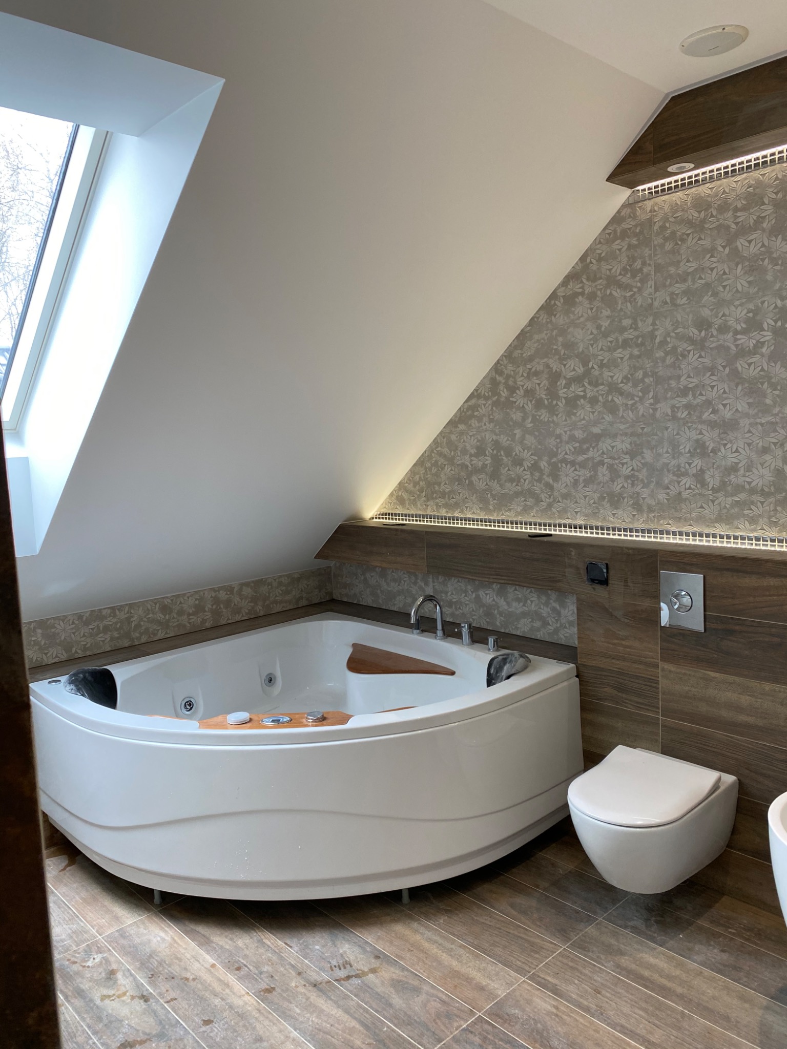 Nowoczesna łazienka na poddaszu z narożną wanną jacuzzi, drewnianym blatem, wiszącą toaletą i płytkami w odcieniach szarości i brązu, oświetlona listwą LED.
