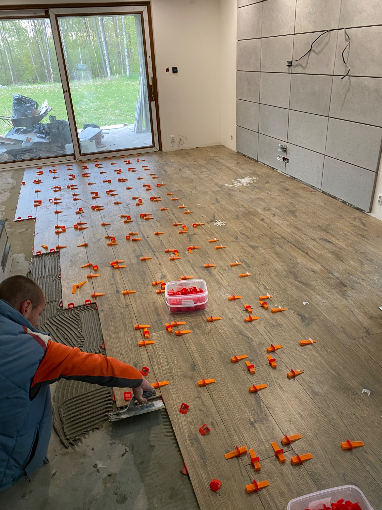 Układanie drewnopodobnych płytek podłogowych z użyciem klipsów poziomujących, widoczny glazurnik nakładający klej na podłoże, w tle okno z widokiem na zieleń i częściowo wykończona ściana z szarych...