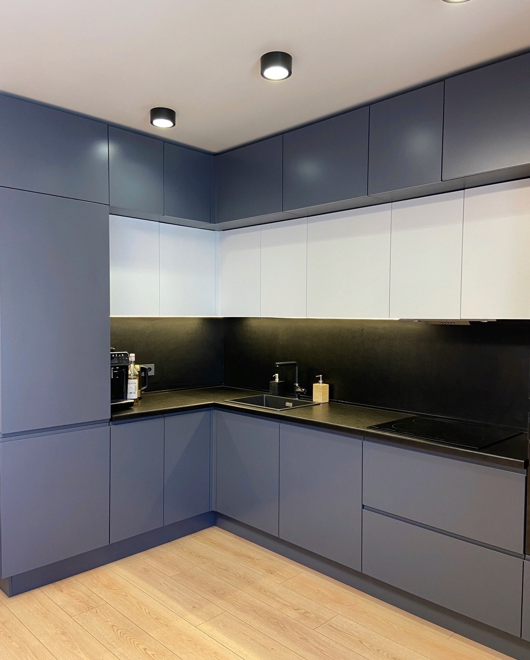 Nowoczesna kuchnia na wymiar w odcieniach szarości i bieli, z minimalistycznym czarnym blatem i drewnianą podłogą, oświetlona punktowymi lampami sufitowymi.