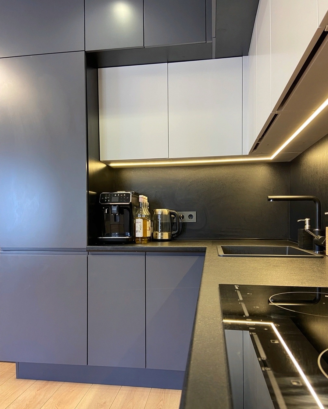 Nowoczesna kuchnia z szarymi szafkami, białymi górnymi szafkami, czarnym blatem i podświetleniem LED pod szafkami wiszącymi; widoczny ekspres do kawy i czajnik na blacie.