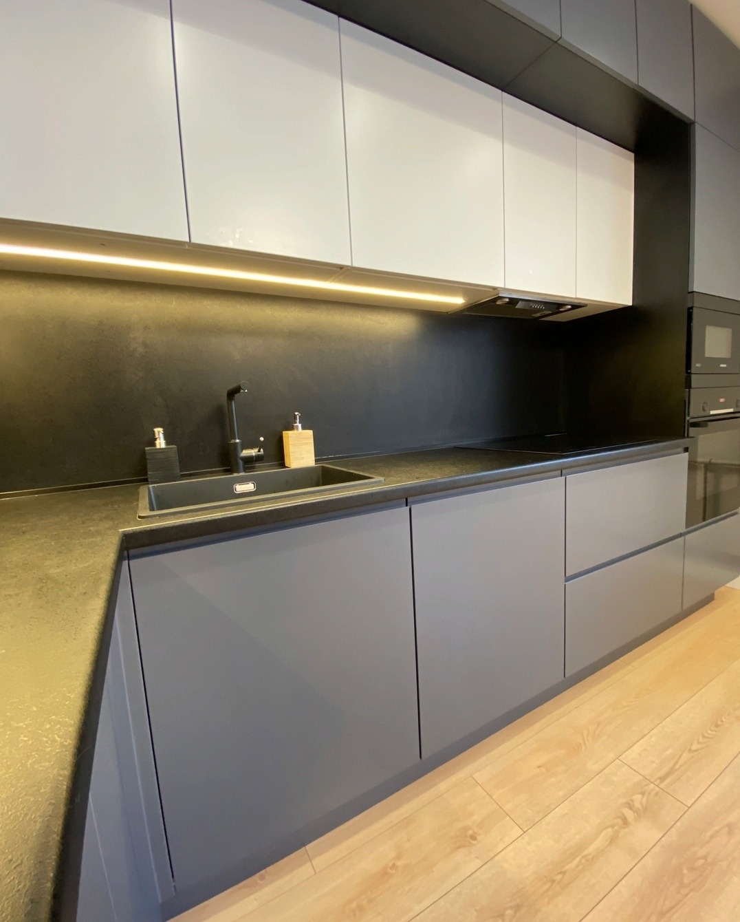 Nowoczesna kuchnia w odcieniach szarości i bieli z podświetleniem LED pod szafkami wiszącymi, widoczny zlewozmywak i bateria w kolorze czarnym.