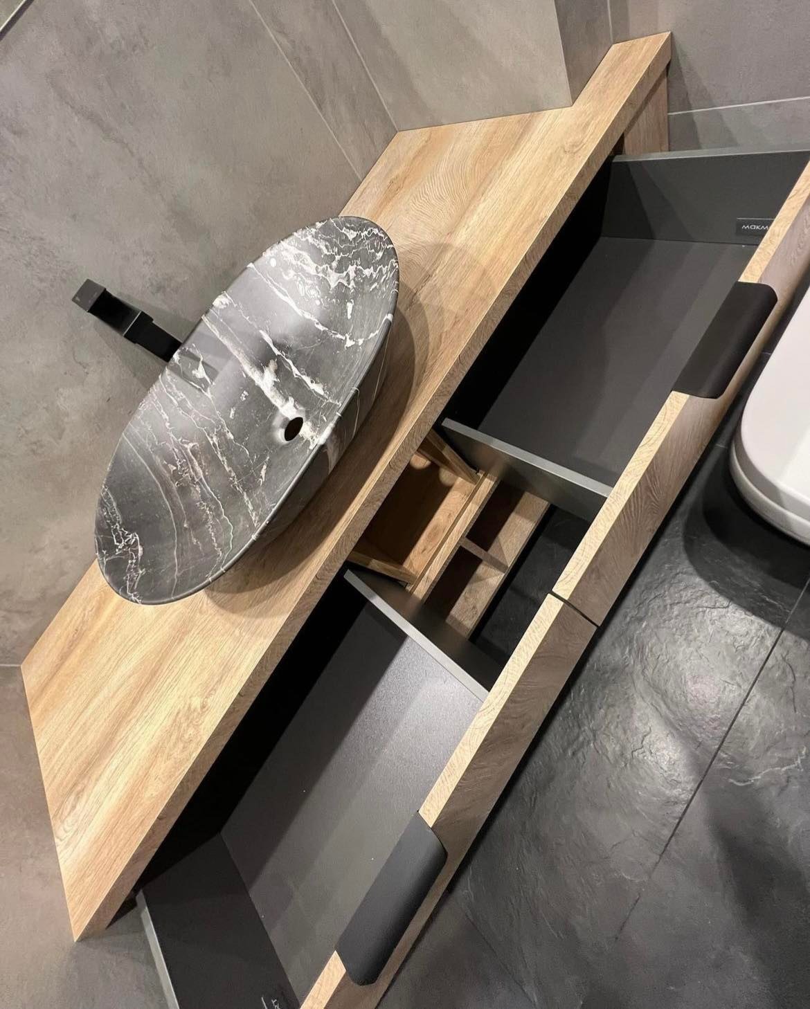 Łazienkowa szafka pod umywalkę z drewna w naturalnym odcieniu, z otwartymi szufladami i umywalką w kolorze szarego marmuru, czarna bateria, szare płytki na podłodze i ścianie.