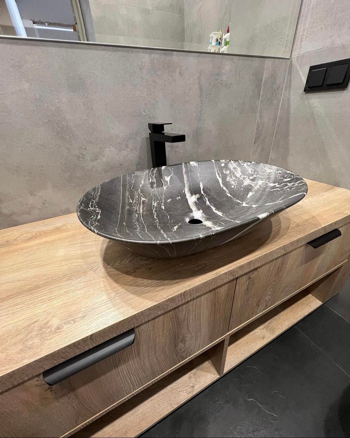 Czarna, marmurkowa umywalka nablatowa na drewnianej szafce z szufladami i czarnymi uchwytami w nowoczesnej łazience.