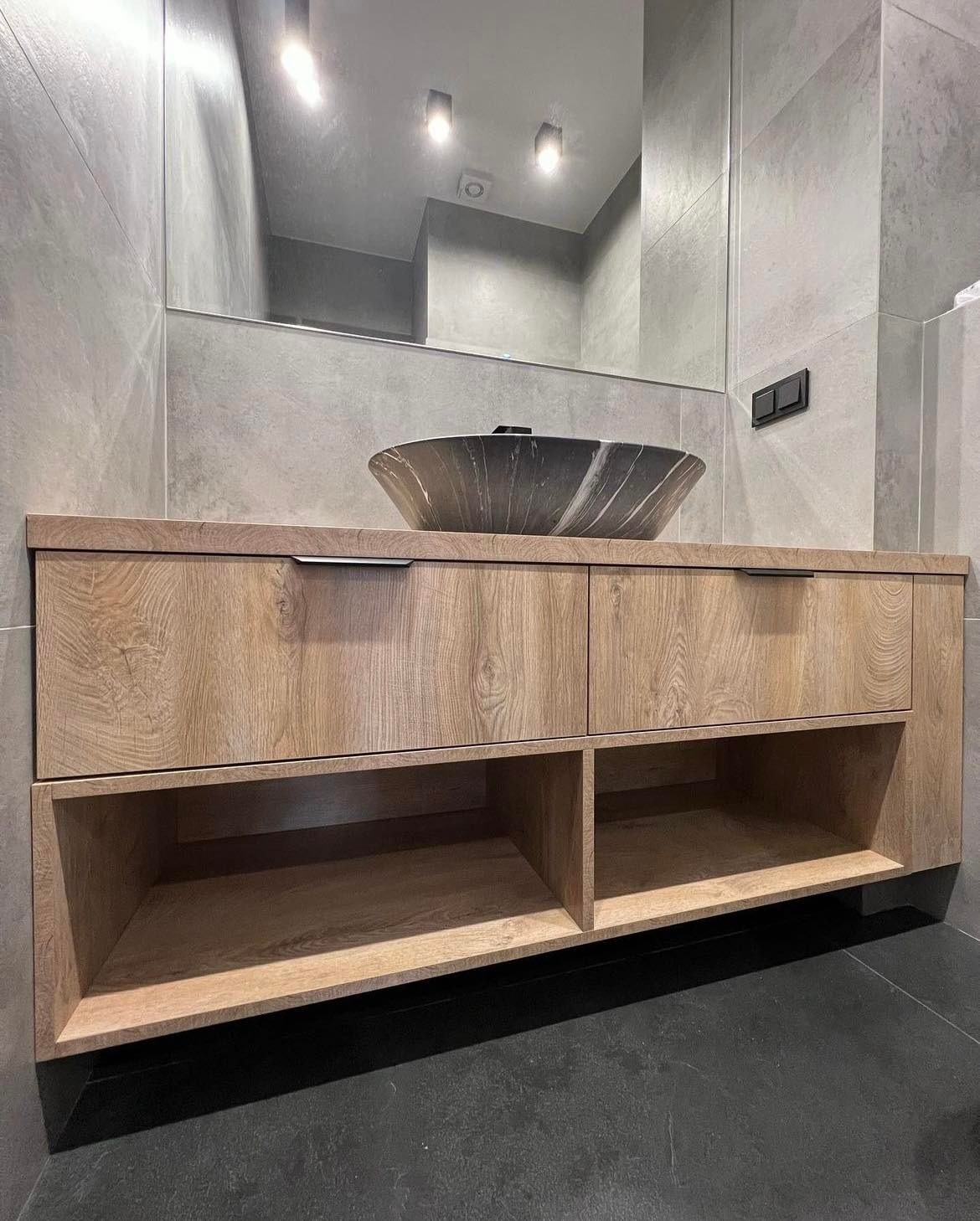 Łazienkowa szafka pod umywalkę z drewnopodobnym frontem, szufladami z minimalistycznymi uchwytami i otwartymi półkami, na tle szarych płytek i czarnej podłogi, z umywalką w odcieniach szarości.