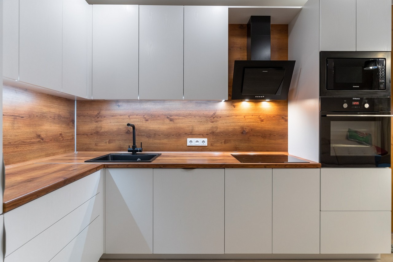 Nowoczesna kuchnia z białymi szafkami, drewnianym blatem i panelem ściennym, czarną armaturą i sprzętem AGD.
