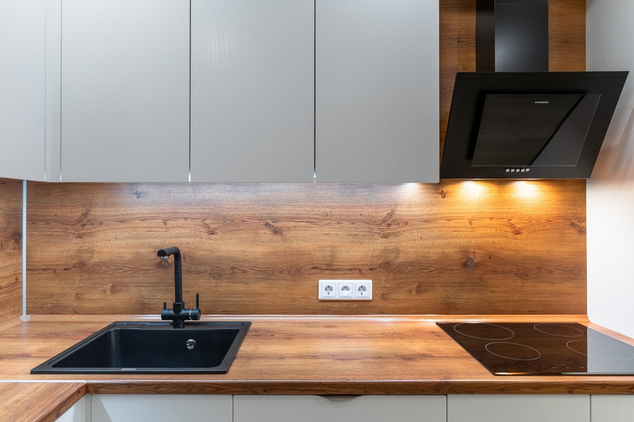 Fragment nowoczesnej kuchni z drewnianym blatem, czarnym zlewem i płytą indukcyjną, oświetlona listwą LED pod szafkami.