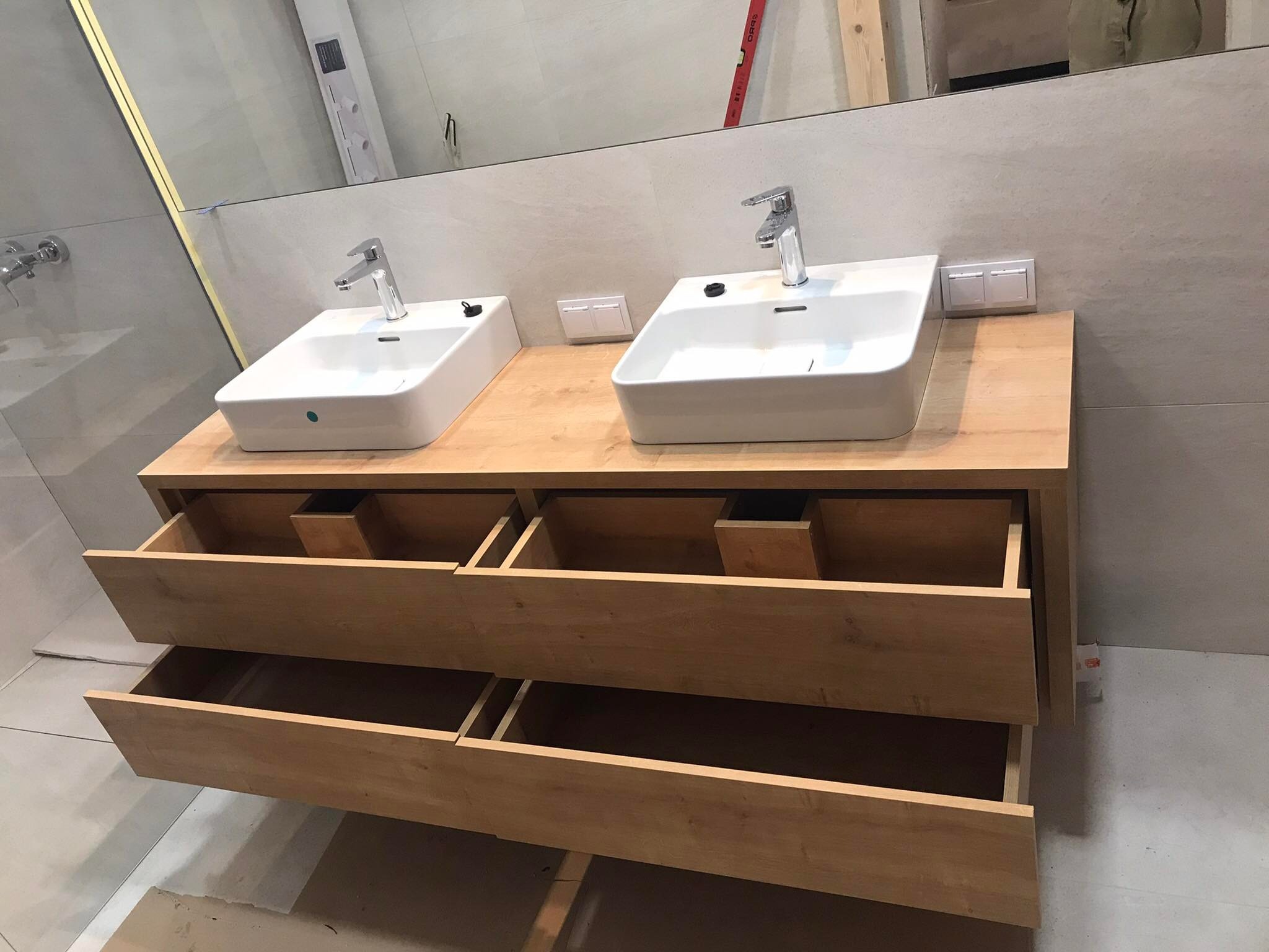 Łazienkowa szafka pod umywalki z drewna z otwartymi szufladami, dwie białe umywalki nablatowe z chromowanymi bateriami, płytki ceramiczne na ścianie i podłodze.