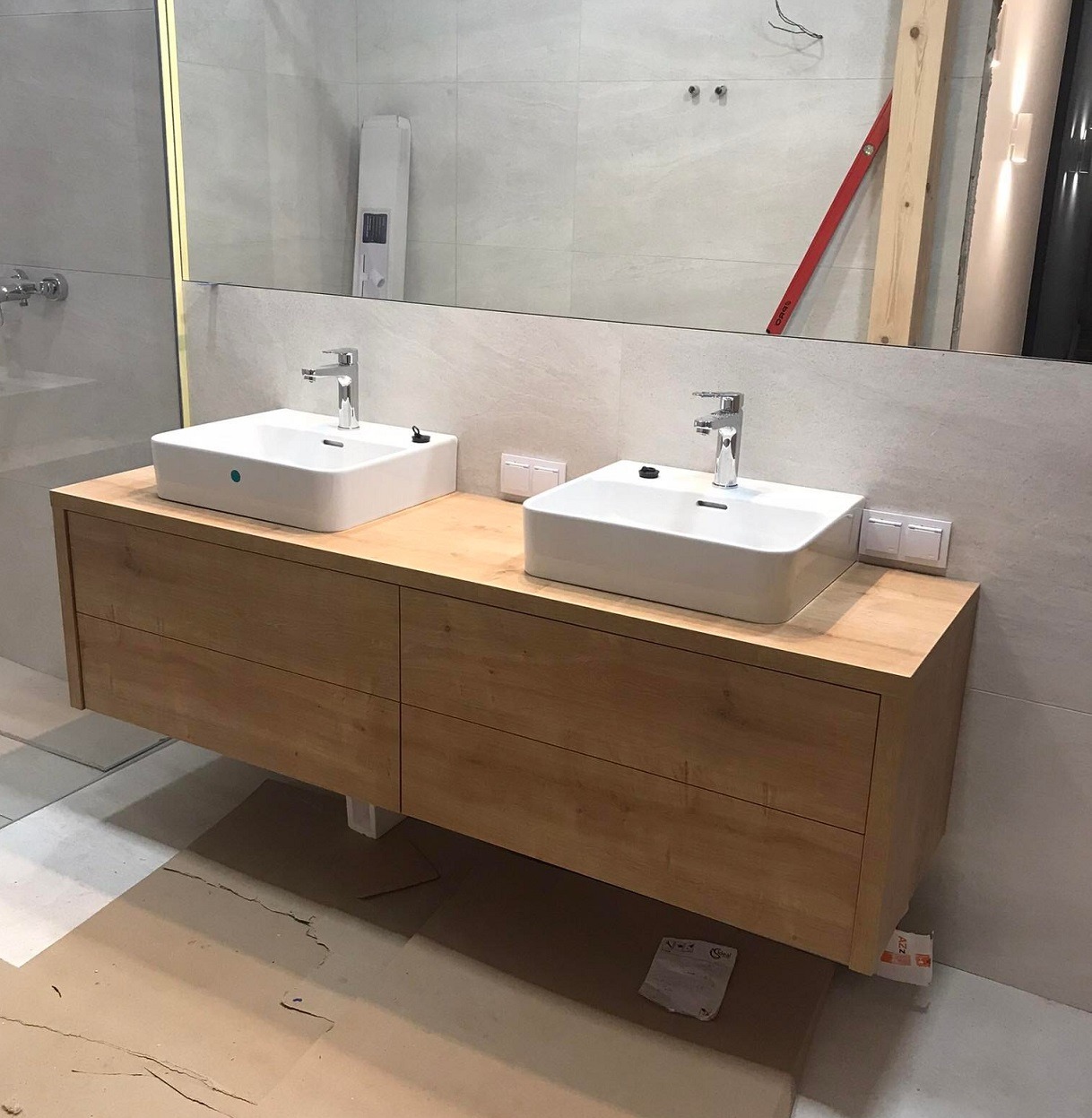 Łazienka w trakcie wykańczania: podwójna umywalka nablatowa o kwadratowym kształcie, osadzona na drewnianej szafce z szufladami, widoczne gniazdka elektryczne i poziomica na ścianie odbijająca się...