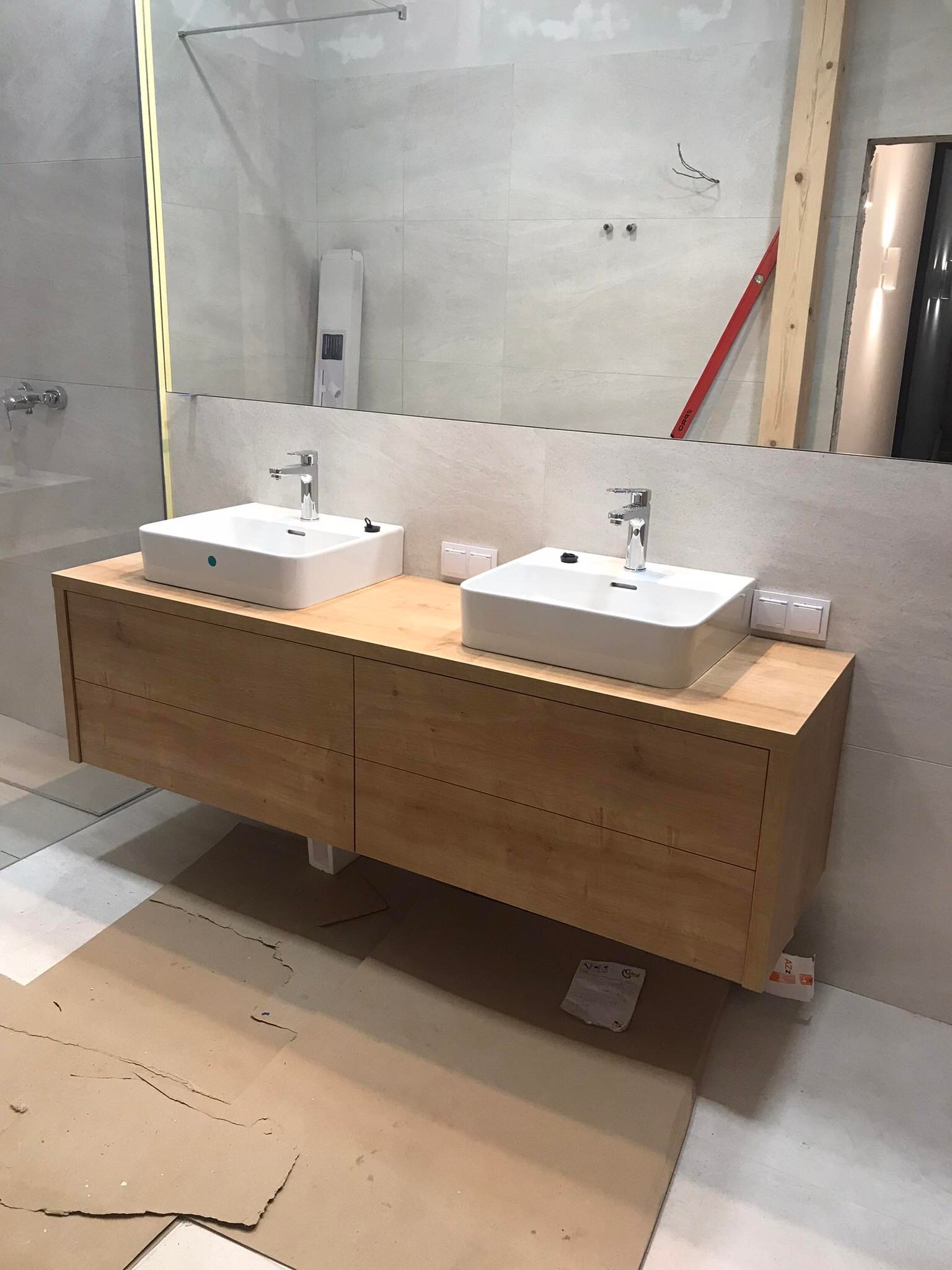 Łazienka w trakcie wykańczania: podwójna umywalka nablatowa na drewnianej szafce z szufladami, duże lustro z oświetleniem, płytki imitujące kamień, widoczne narzędzia i zabezpieczenia podłogi.