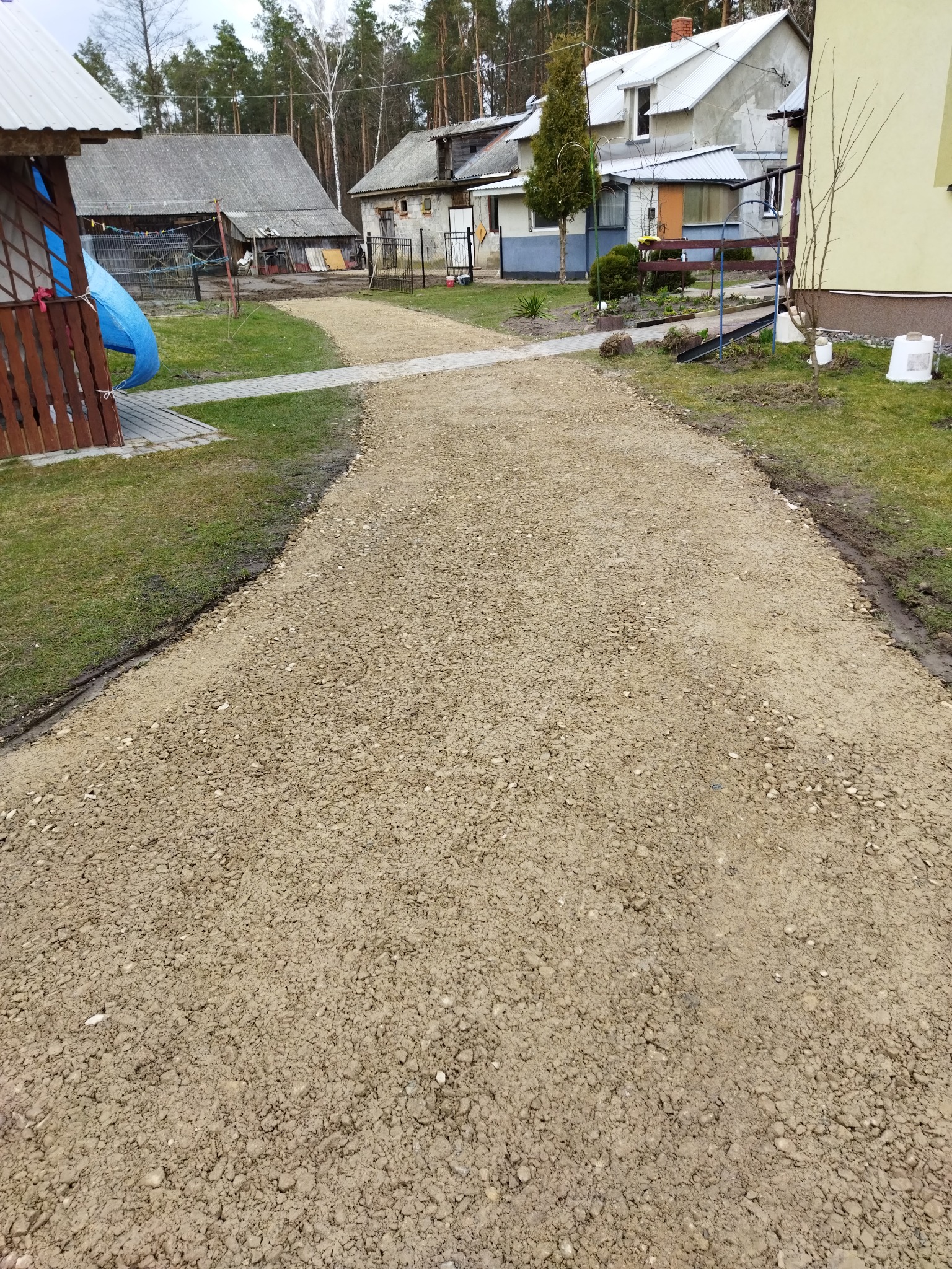 Nierówna, szutrowa droga gruntowa prowadząca w kierunku zabudowań wiejskich, z widocznymi śladami prac ziemnych po bokach i fragmentem ułożonej kostki po lewej stronie.