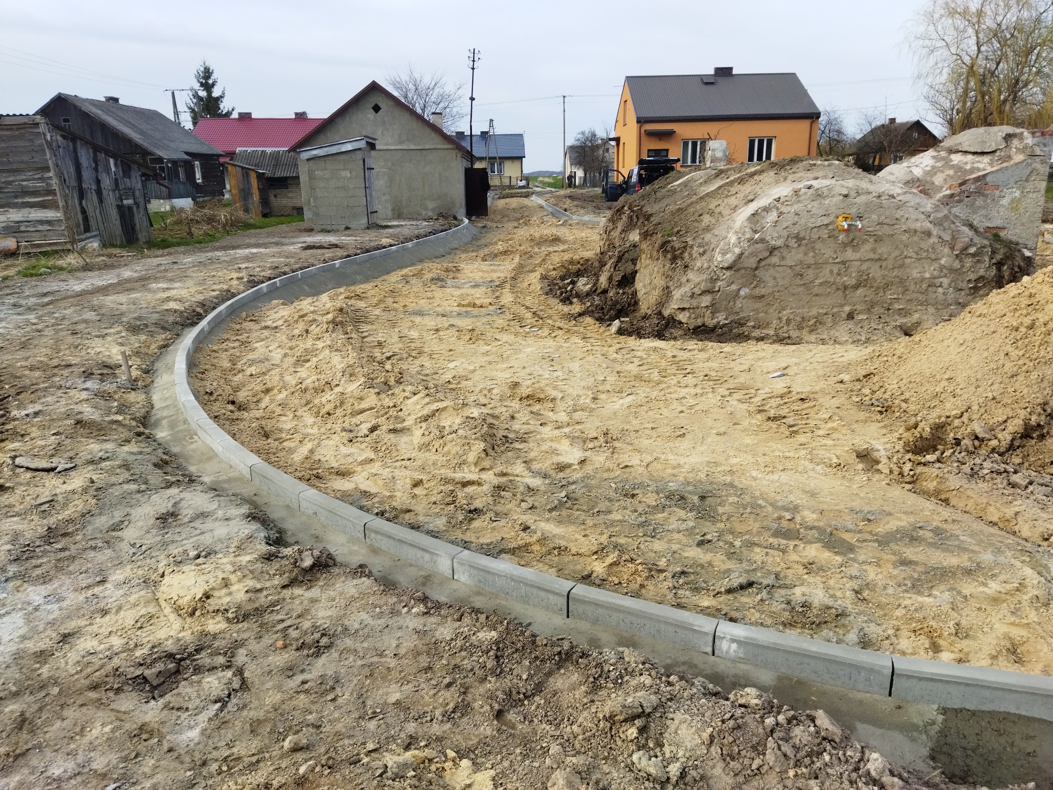 Układanie obrzeży betonowych na piaszczystym podłożu wzdłuż drogi gruntowej, w tle zabudowa wiejska i hałda ziemi po wykopach.