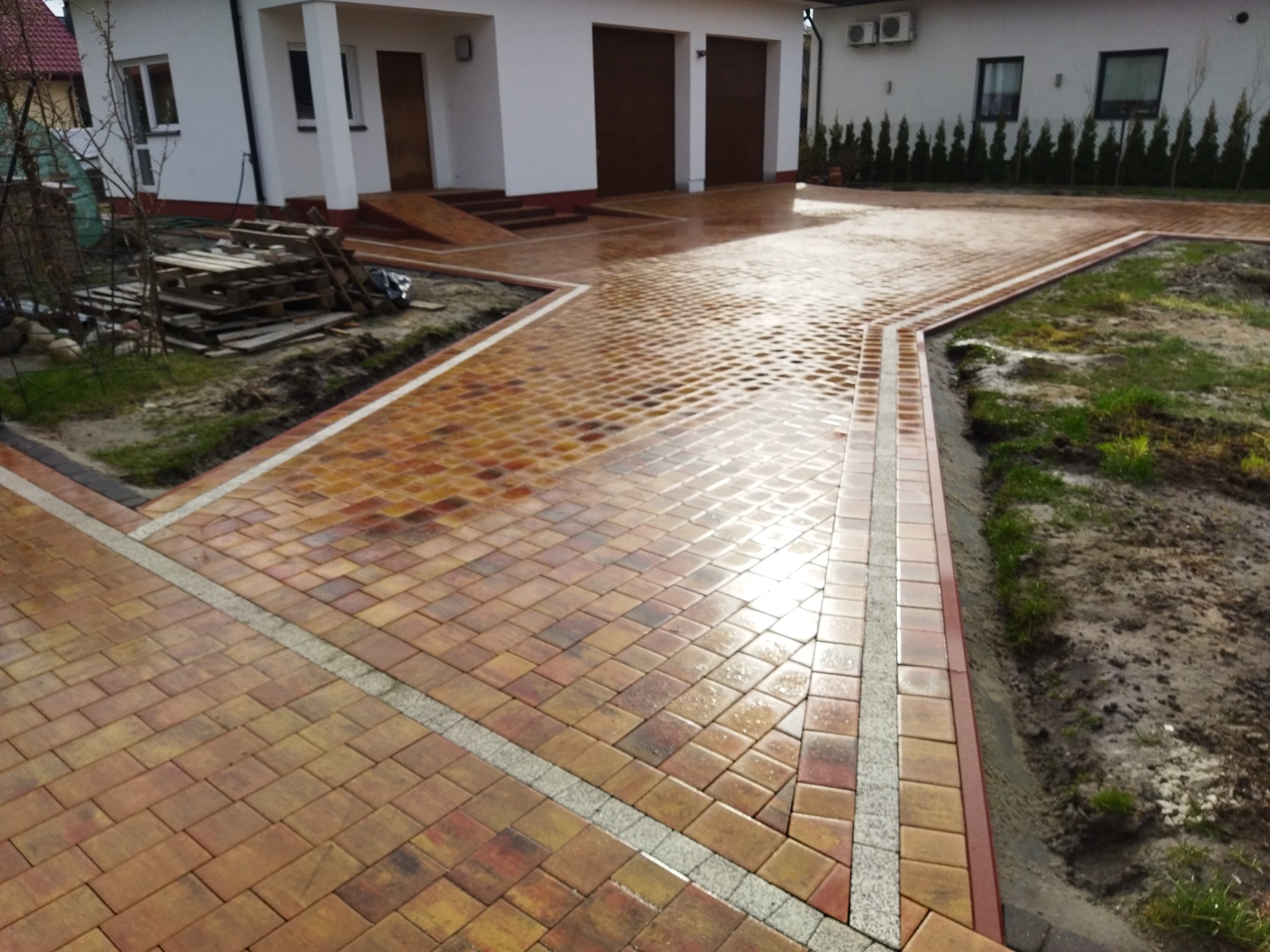 Nowo ułożona, wilgotna kostka brukowa w odcieniach brązu i czerwieni, z jasnoszarymi obramowaniami, tworząca wzór na podjeździe przed domem z garażem dwustanowiskowym; w tle widoczne palety...