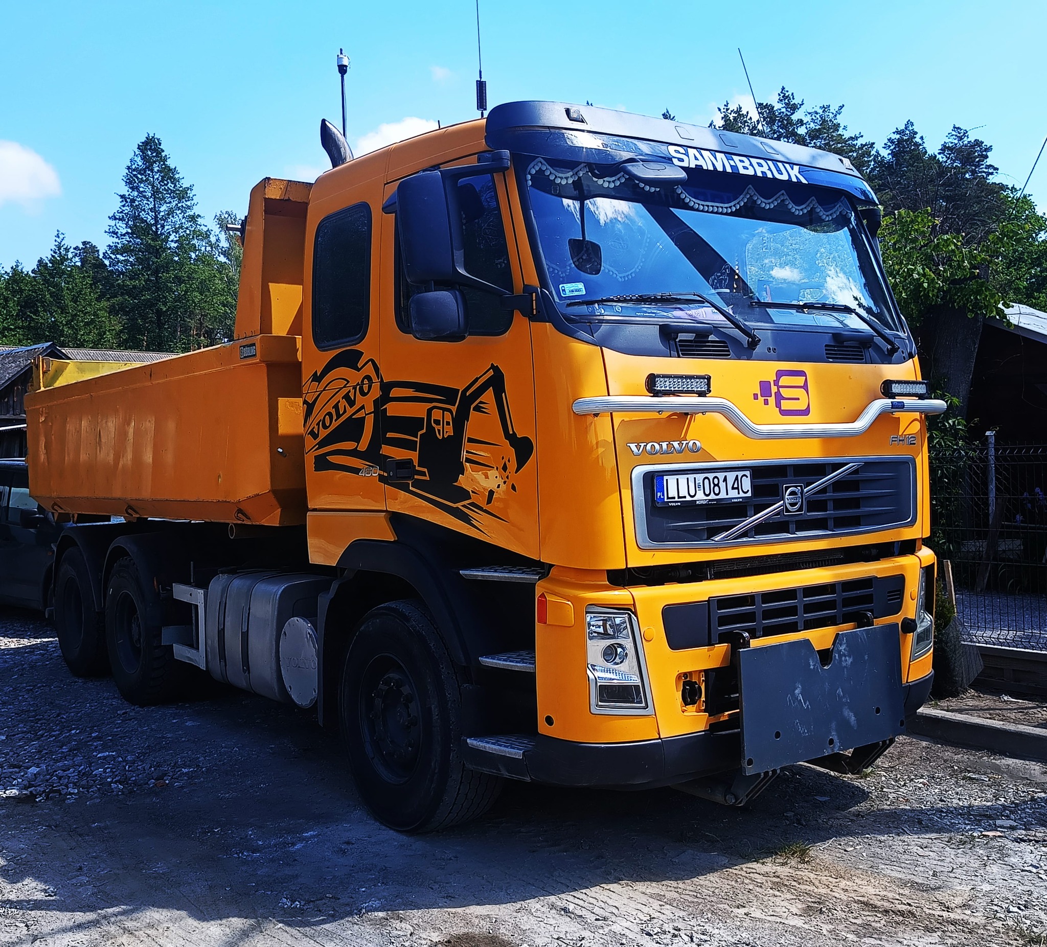 Żółta wywrotka Volvo z naklejką przedstawiającą koparkę, zaparkowana na żwirowym podłożu, z widocznym fragmentem kabiny i skrzyni ładunkowej.