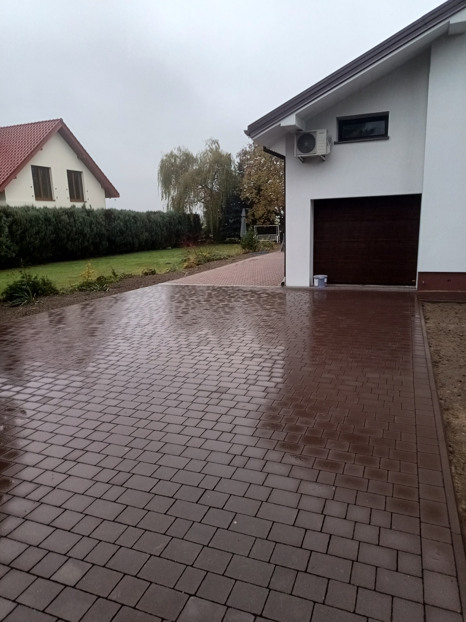 Świeżo ułożona, mokra kostka brukowa w odcieniach brązu prowadząca do garażu w nowoczesnym domu z białą elewacją, widoczny fragment ogrodu i sąsiedniego domu w tle.