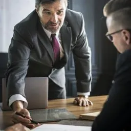 Mężczyzna w garniturze pochylony nad stołem, wskazujący długopisem na dokumenty podczas spotkania z klientem. Widoczny laptop w tle.