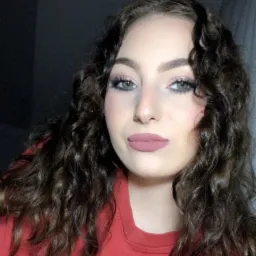 Portret młodej kobiety z delikatnym makijażem oczu i matowymi ustami w kolorze dusty rose, z ciemnymi kręconymi włosami, ubrana w czerwoną bluzę z logo Nike, na ciemnym tle.