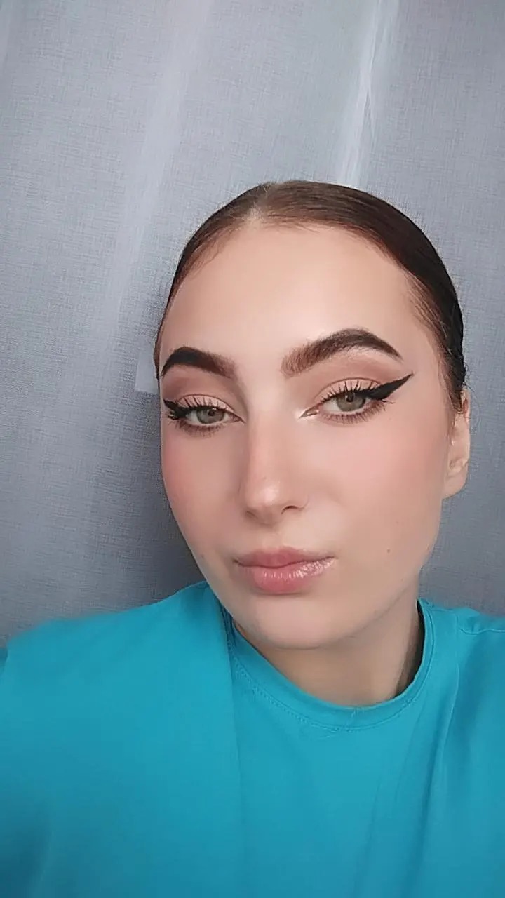 Portret kobiety z precyzyjnym makijażem oczu, wyraźne kreski eyelinerem i delikatny makijaż ust, na tle szarej zasłony.