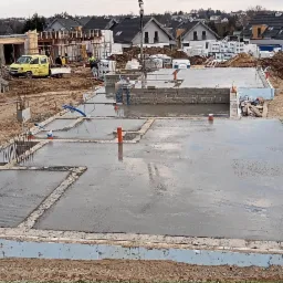 Wylany, mokry beton na fundamencie domu jednorodzinnego w trakcie budowy, widoczne wystające pręty zbrojeniowe, izolacja fundamentu, na drugim planie betonowóz i budowa domu z białych bloczków.