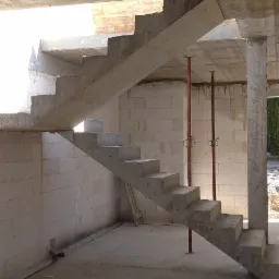 Surowe schody z betonu w trakcie budowy, wsparte tymczasowymi podporami, widoczne ściany z bloczk&oacute;w i surowy betonowy strop.