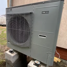 Szara pompa ciepła NIBE AMS10, zamontowana na betonowych bloczkach, widoczna z boku na tle ściany budynku, z elementami instalacji i otaczającym terenem.