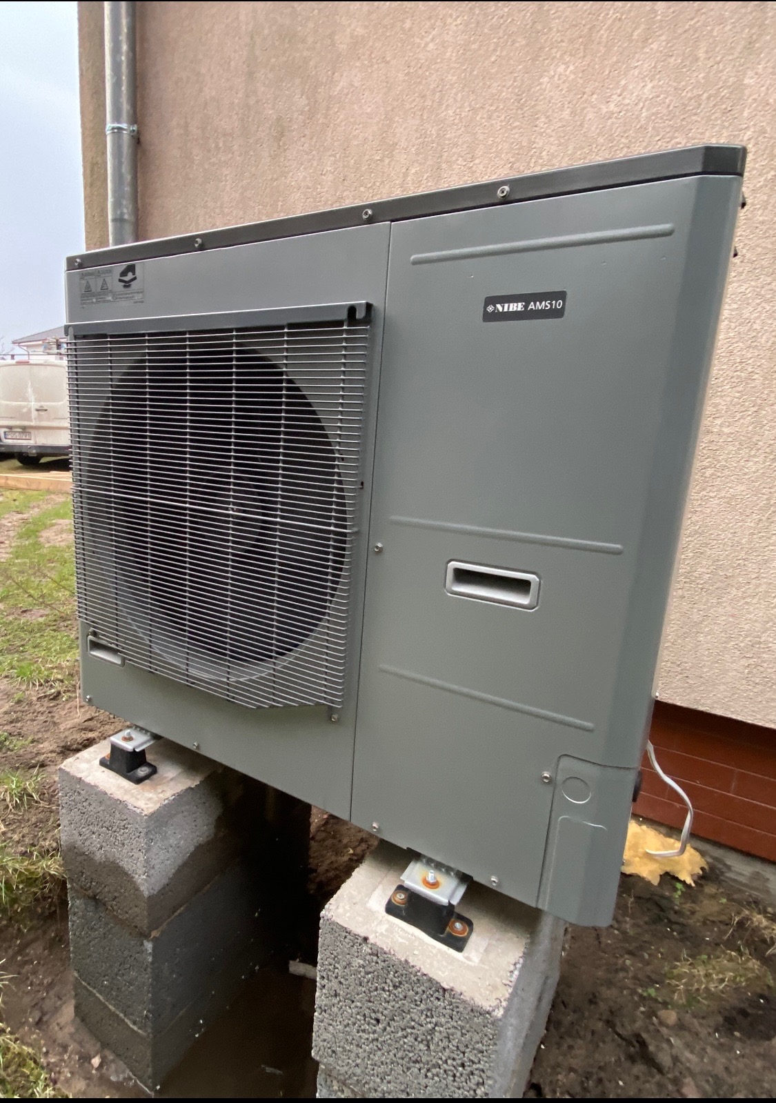 Szara pompa ciepła NIBE AMS10, zamontowana na betonowych bloczkach, widoczna z boku na tle ściany budynku, z elementami instalacji i otaczającym terenem.