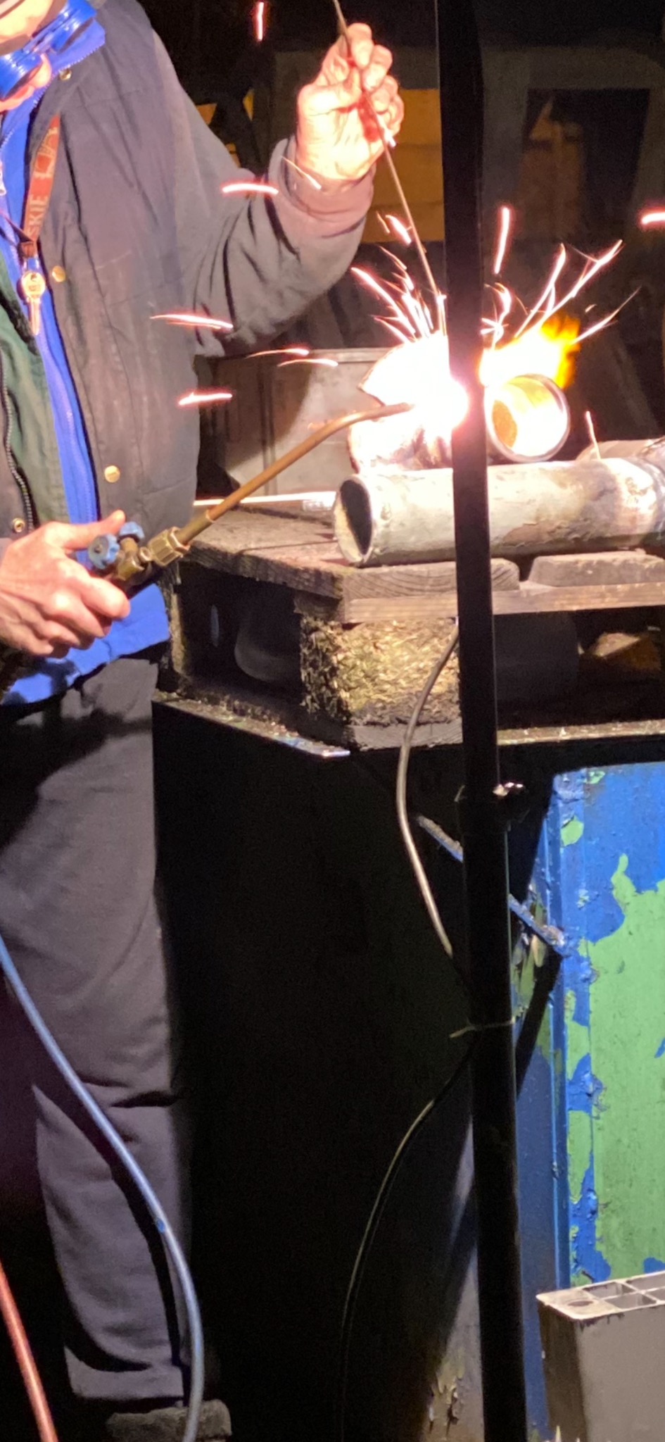 Spawanie rury gazowej palnikiem acetylenowo-tlenowym, widoczne iskry i płomień, rura na prowizorycznym stanowisku roboczym, pracownik w ubraniu roboczym.