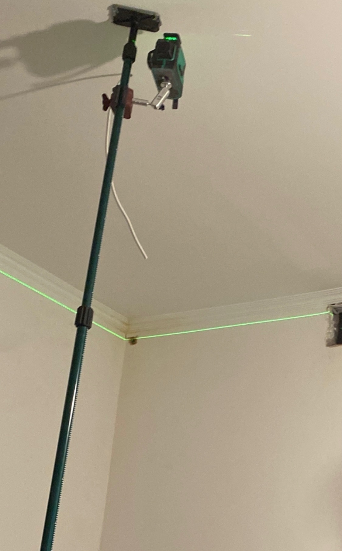 Poziomica laserowa ustawiona na wysięgniku, wyświetlająca zielone linie na ścianie i suficie podczas prac instalacyjnych.