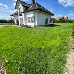 Nowoczesny dom jednorodzinny z białymi ścianami i ciemną dachówką otoczony świeżo założonym trawnikiem, widoczne ślady po pracach ziemnych na obrzeżach, błękitne niebo z chmurami.