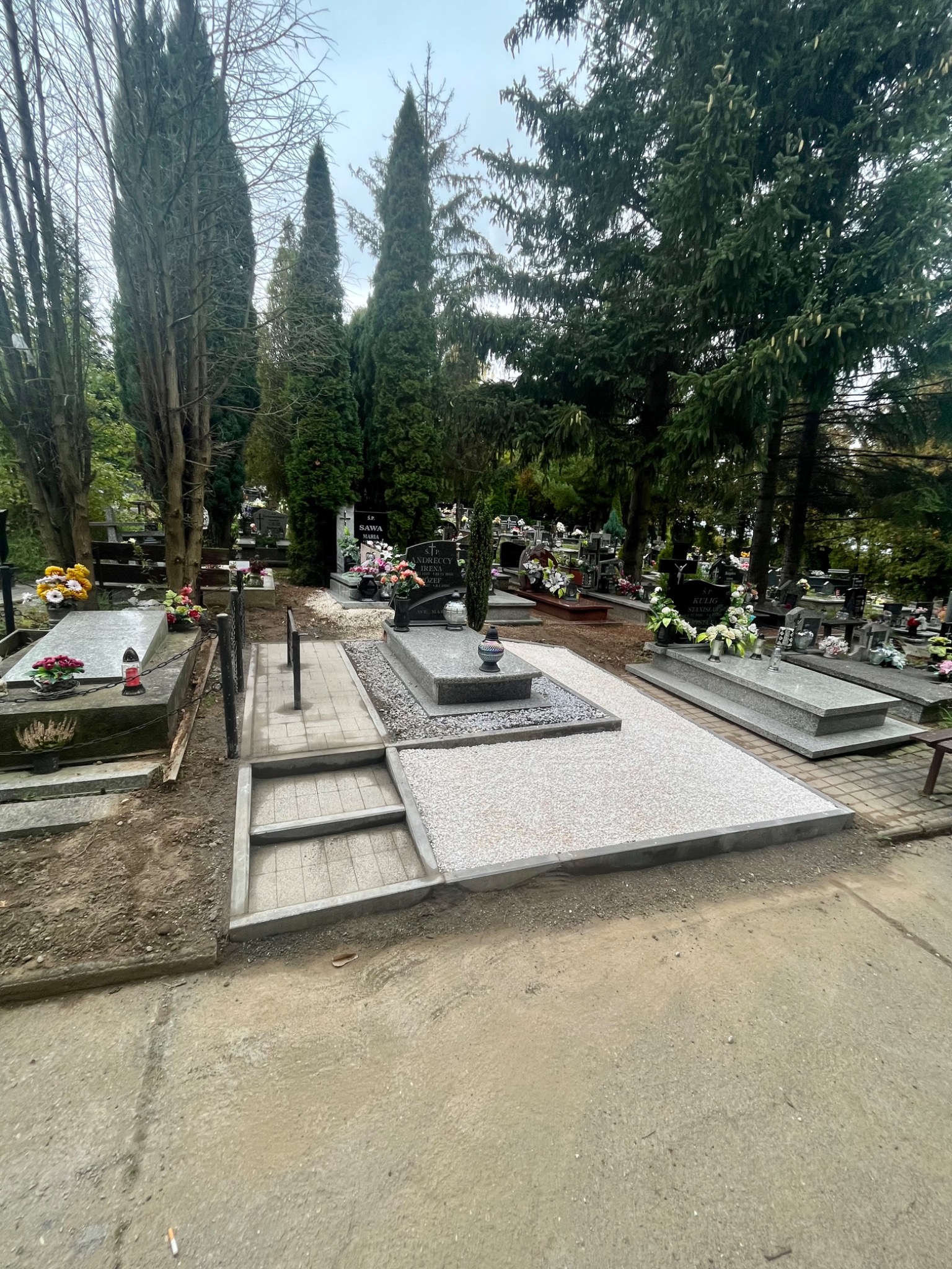 Nowo wykończony nagrobek z jasnoszarą, żwirową powierzchnią, schodami z kostki brukowej i otaczającą zielenią cmentarza.