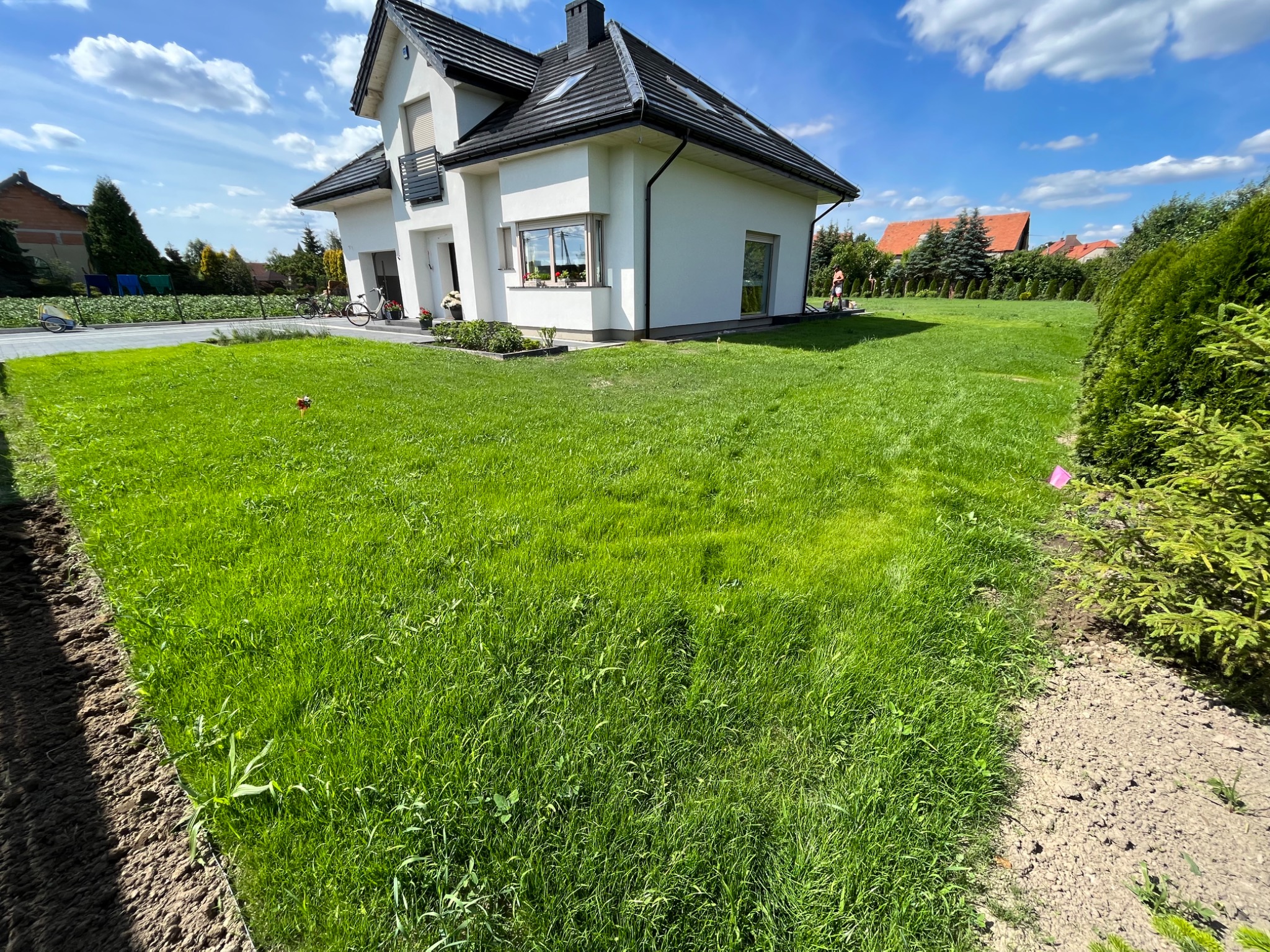 Nowoczesny dom jednorodzinny z białymi ścianami i ciemną dachówką otoczony świeżo założonym trawnikiem, widoczne ślady po pracach ziemnych na obrzeżach, błękitne niebo z chmurami.