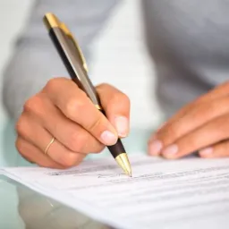 Dłoń kobiety z pierścionkiem podpisuje dokument eleganckim długopisem na szklanym biurku.
