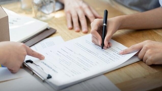 Ręka podpisuje czarnym długopisem umowę na podkładce, druga ręka wskazuje palcem na dokument, drewniany stół w tle.