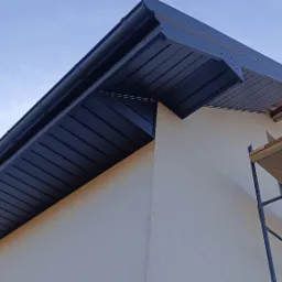 Nowoczesne, grafitowe podbitki dachowe z PVC, zamontowane na budynku o jasnej elewacji. Widoczne rusztowanie. Ujęcie z dołu, podkreślające estetykę wykończenia.