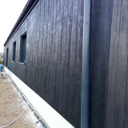 BUDROB - Ciemna, drewnopodobna elewacja budynku z widocznymi oknami i rurą spustową. U dołu biały cokół. Ujęcie z boku, podkreślające fakturę i kolorystykę.