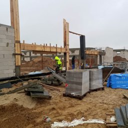 BUDROB - Budowa domu w Augustowie: wznoszenie konstrukcji szkieletowej z drewna i bloczków betonowych. Widoczny pracownik w odblaskowej kamizelce i materiały budowlane.