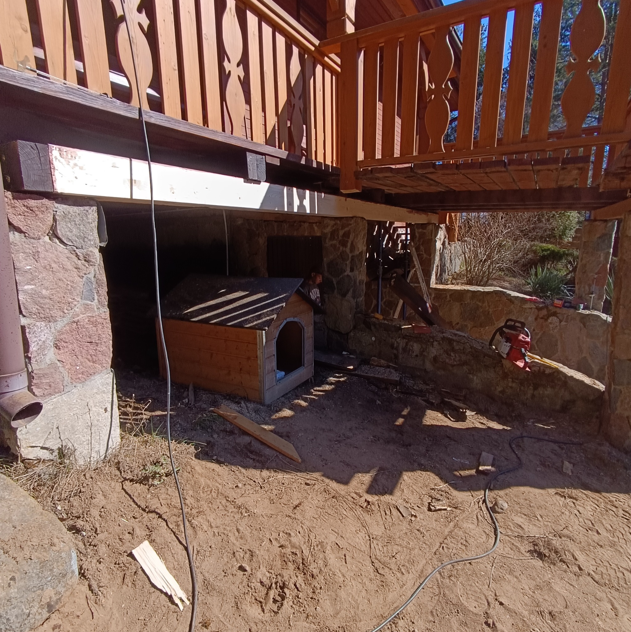 Drewniany domek dla psa z zadaszeniem, umieszczony pod podwyższonym tarasem domu z kamienną podmurówką. W tle widoczny mężczyzna i narzędzia.