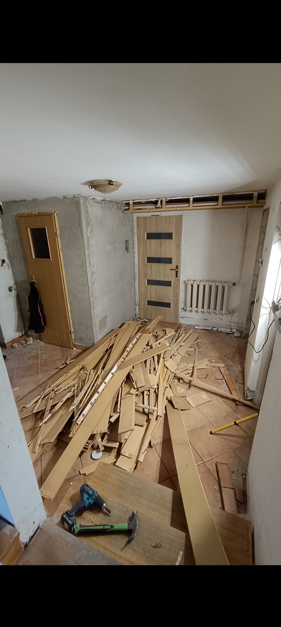 Demontaż podłogi: sterta paneli, wiertarka i młotek w pokoju z dwoma drzwiami i grzejnikiem. Widoczna konstrukcja podwieszanego sufitu. Etap prac remontowych.