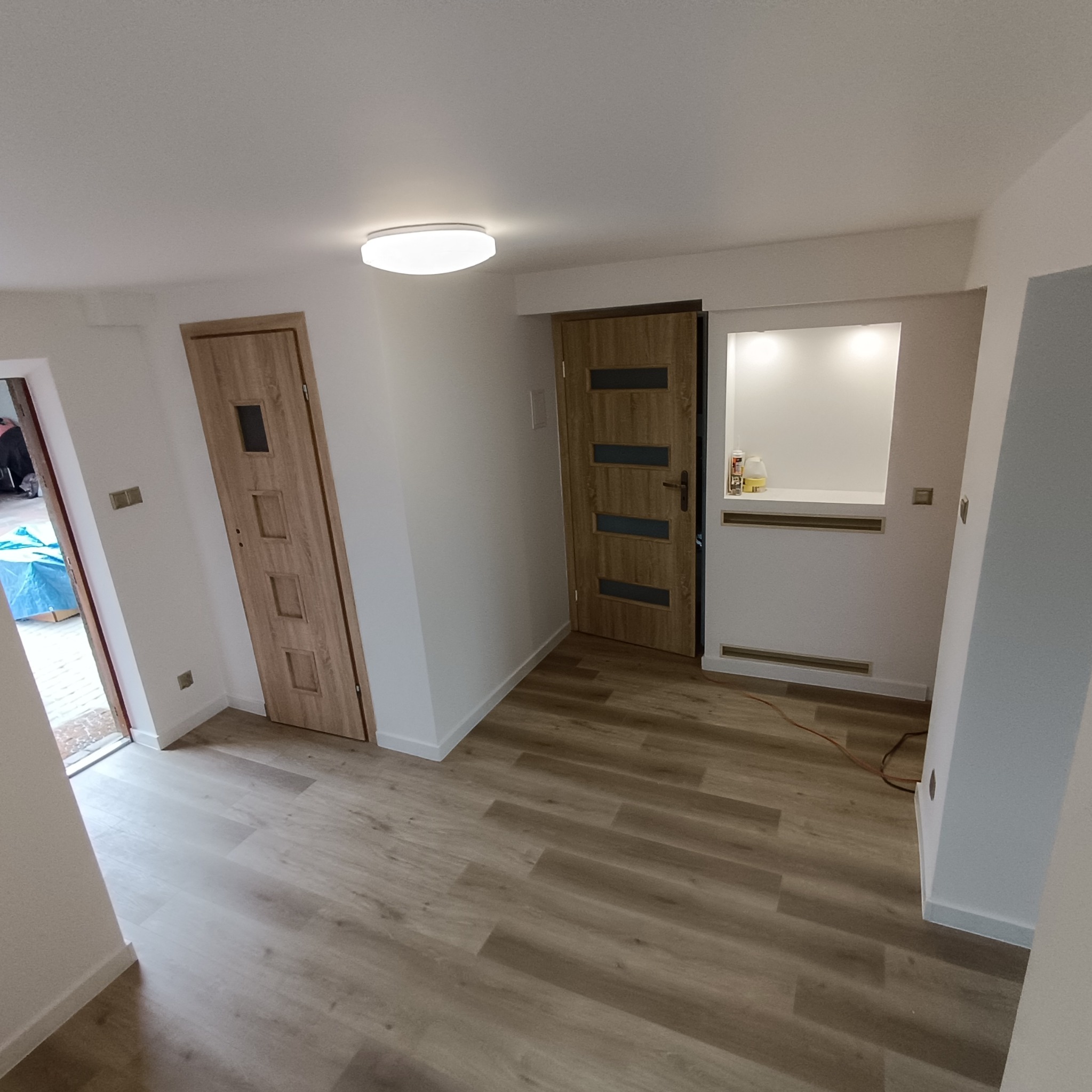 Jasne panele podłogowe w wyremontowanym wnętrzu z dwiema drewnianymi drzwiami i wnęką z oświetleniem. Widoczne listwy przypodłogowe i gładkie, białe ściany.