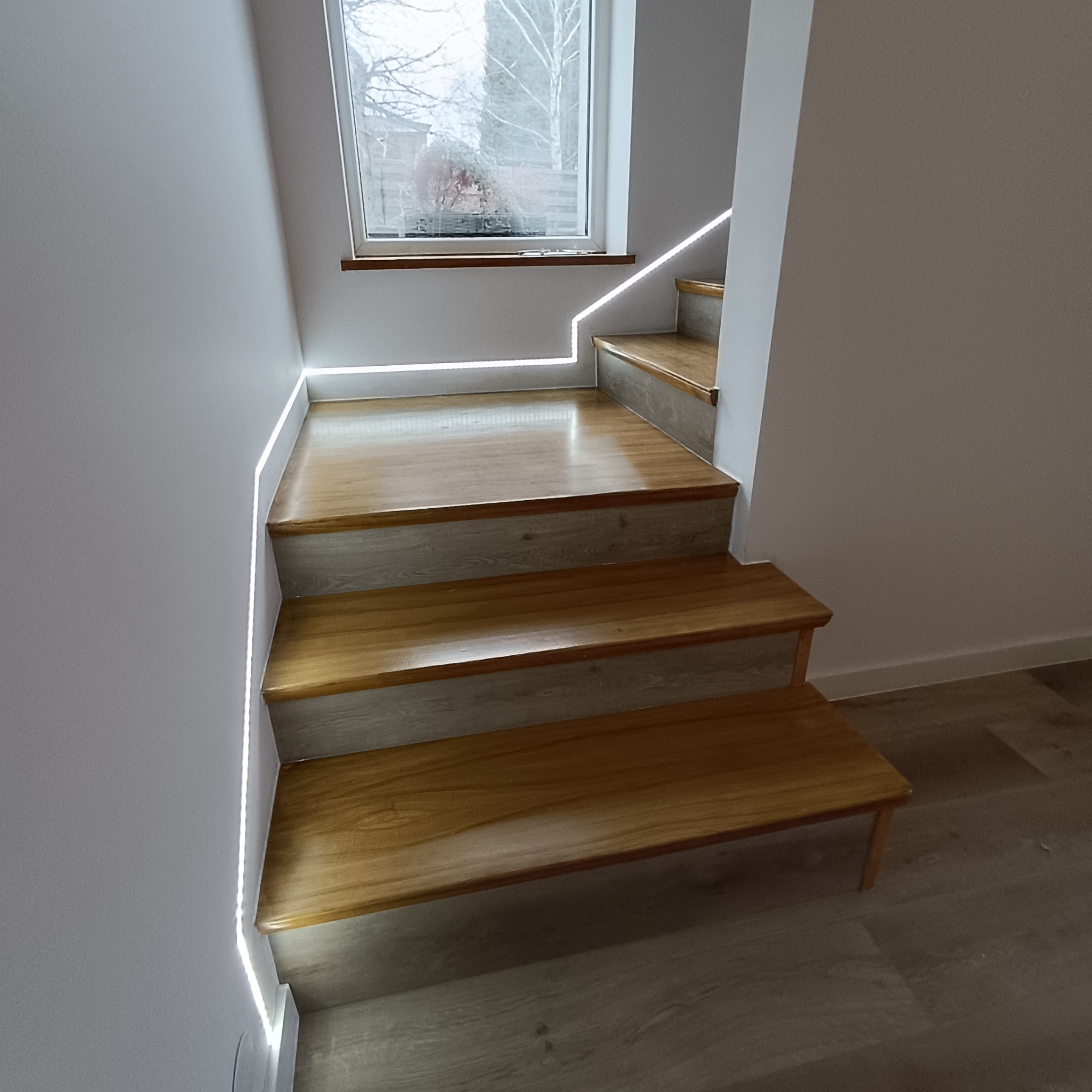 Drewniane schody z subtelnym oświetleniem LED wzdłuż ściany, prowadzące w górę. Nowoczesny design z widokiem z okna. Jasne, stonowane kolory.