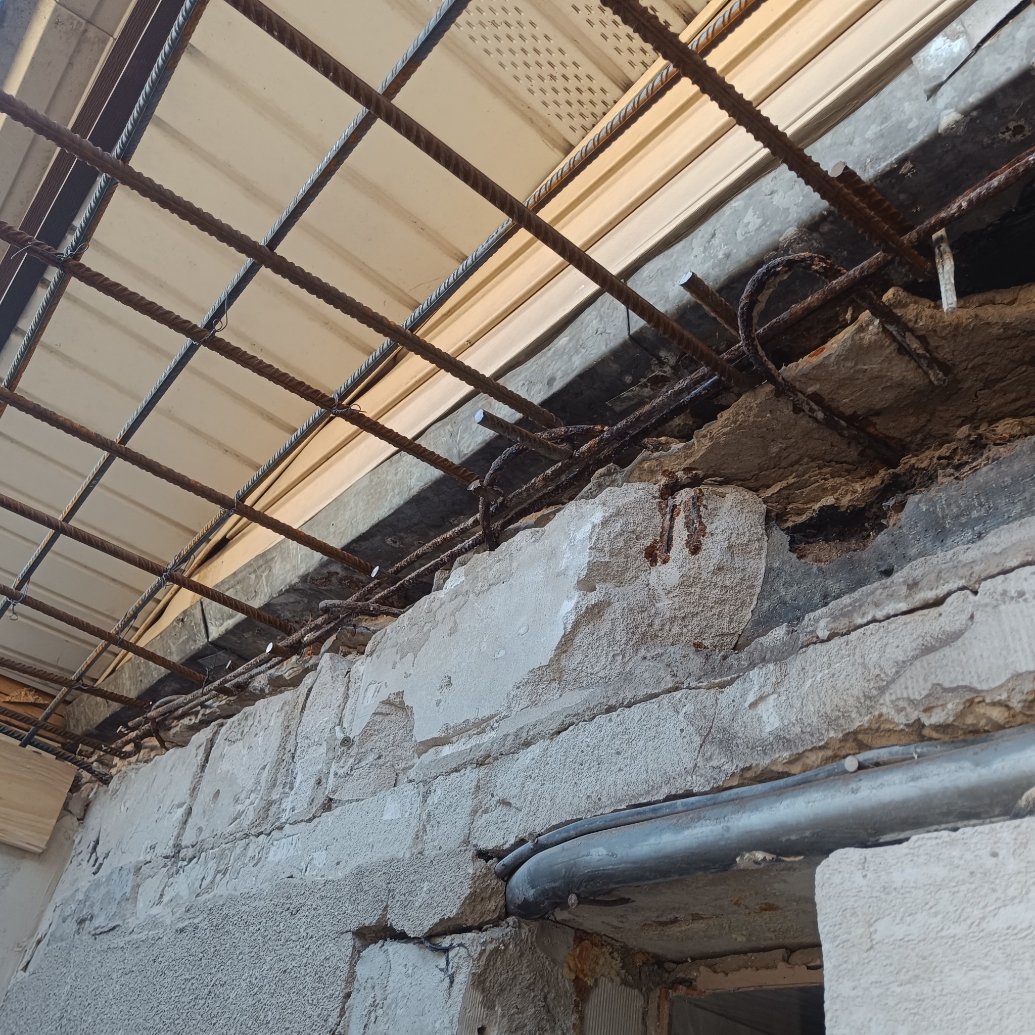 Zbrojenie stropu z siatki stalowej, widoczne uszkodzenia betonu i odsłonięta konstrukcja. Fragment ściany z widocznymi pęknięciami i instalacją elektryczną.