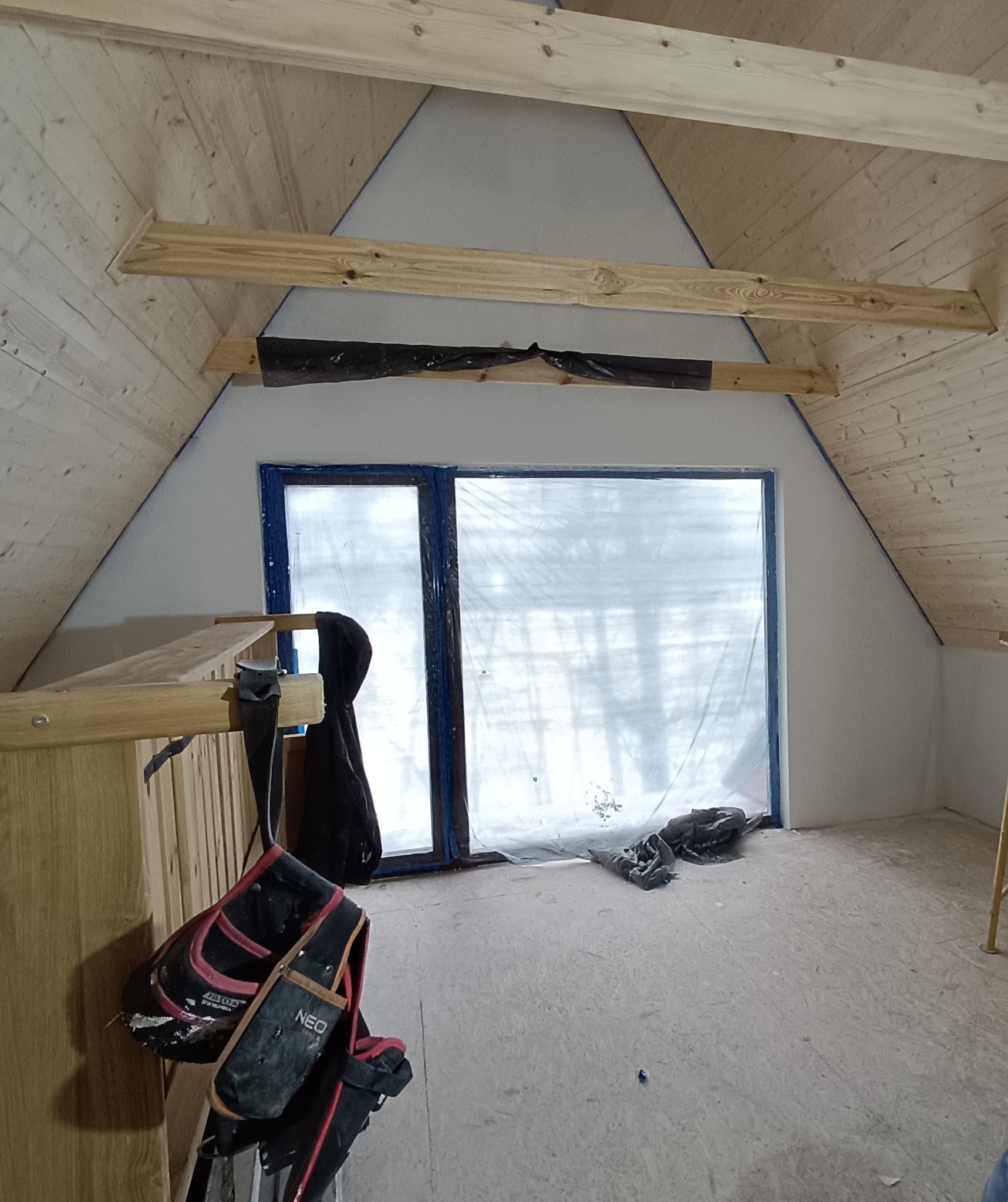 Poddasze w trakcie adaptacji: surowe drewno, okno zabezpieczone folią, widoczne narzędzia. Etap prac przygotowawczych do wykończenia wnętrza.