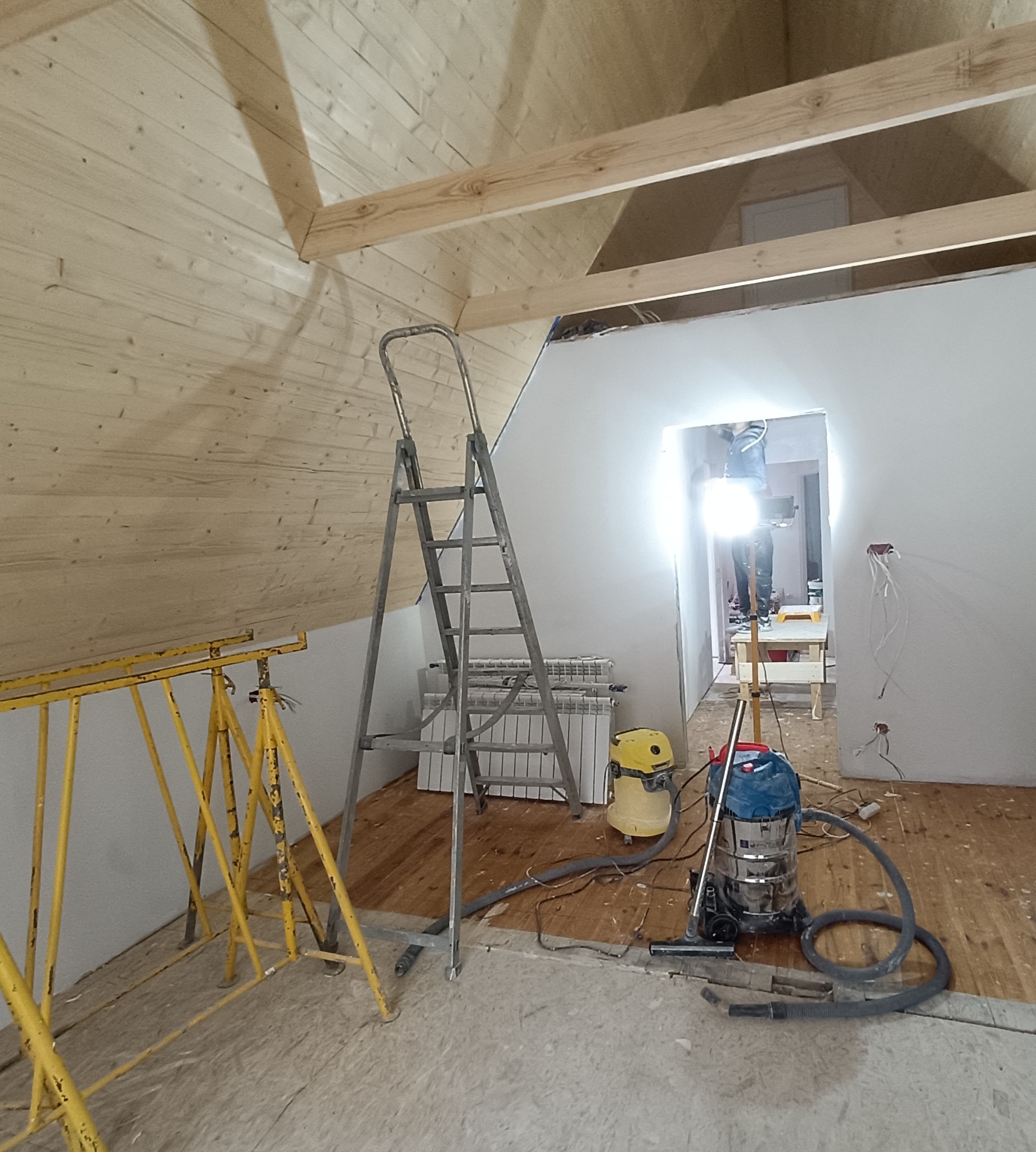 Poddasze w trakcie prac wykończeniowych: drewniana boazeria na suficie, drabina, odkurzacze, widoczny etap prac remontowych. Surowy stan podłogi, instalacja elektryczna.