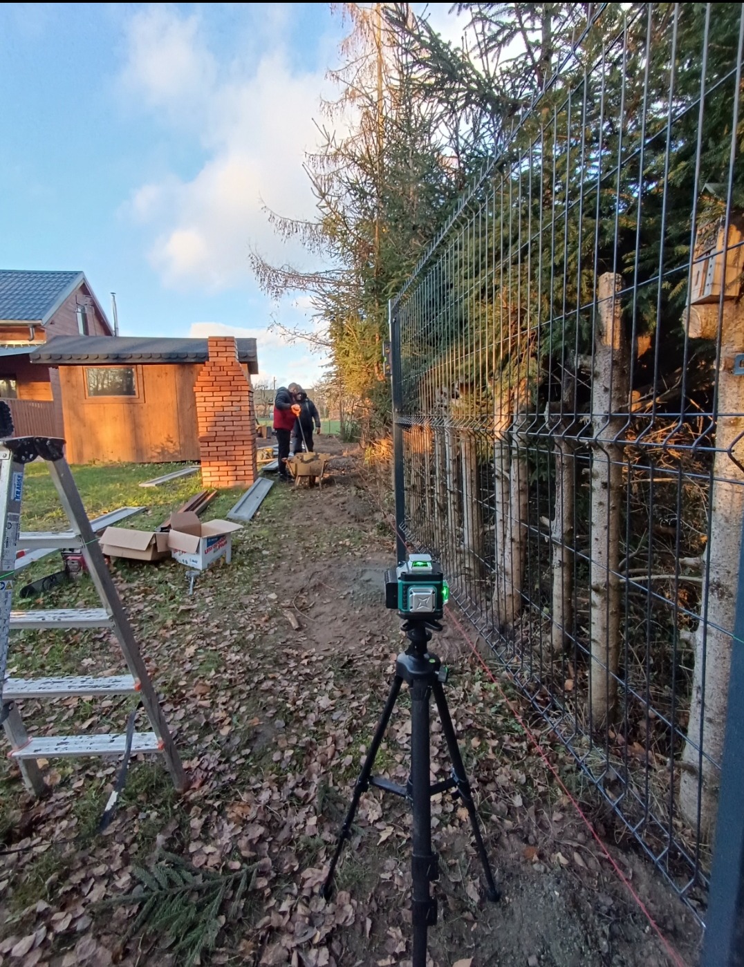 Montaż ogrodzenia panelowego z użyciem poziomicy laserowej. Widoczny fragment działki, drabina i pracownicy w tle.