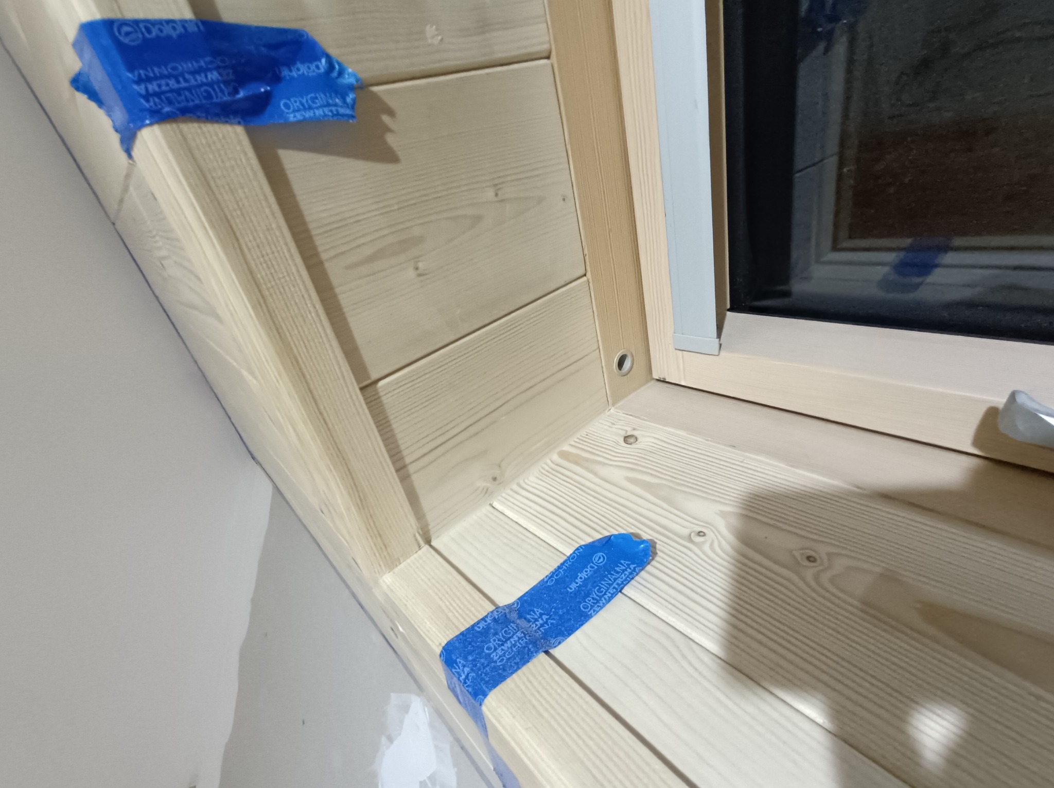 Drewniane wykończenie okna z zabezpieczeniem folią malarską. Widoczny fragment ściany. Ujęcie z bliska, podkreślające detale i precyzję wykonania. Jasne drewno.